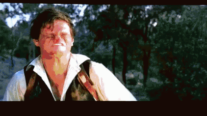 a man in a white shirt and vest is making a funny face while standing in a forest .