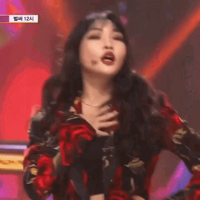 a woman in a red and black floral dress is standing on a stage with her hands on her chest .