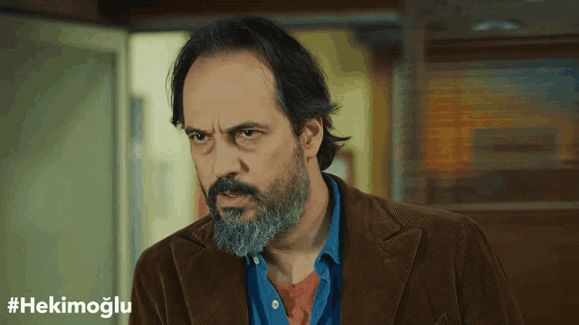 a man with a beard wearing a brown jacket has #hekimoglu written on the bottom