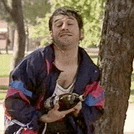 a man in a blue and red jacket is leaning against a tree holding a bottle .