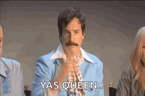 a man with a mustache is sitting in front of a group of people and saying yas queen