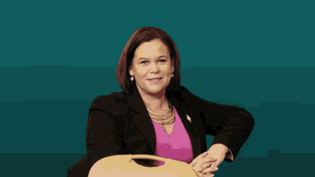 a woman wearing a black jacket and a pink shirt is sitting in front of a blue background
