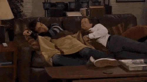 two men are sleeping on a couch in a living room with a remote control in their hand .