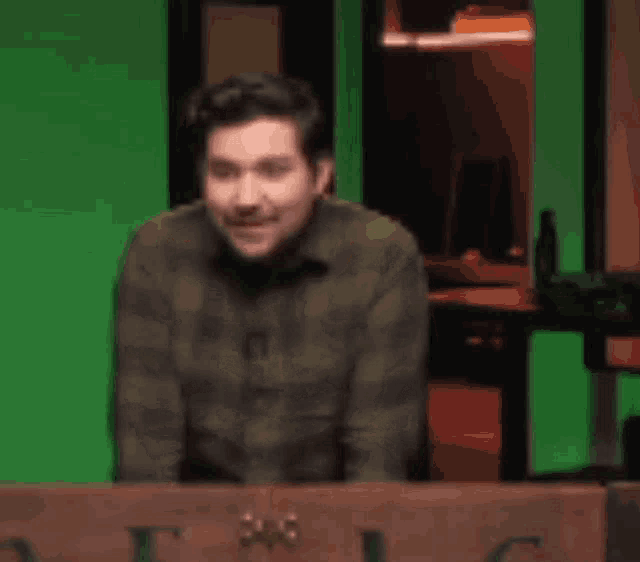 a man in a plaid shirt is sitting at a table with a green background .