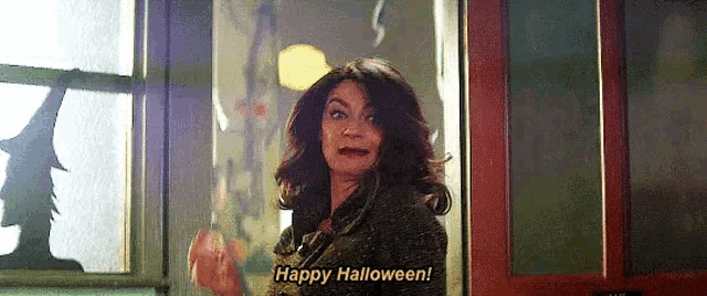 a woman is standing in a doorway with a silhouette of a witch on the window and says `` happy halloween '' .
