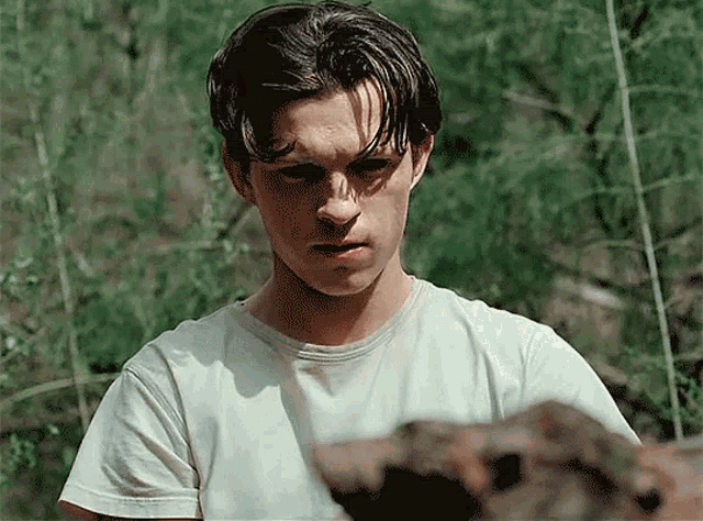a young man in a white t-shirt is standing in a field looking at something .