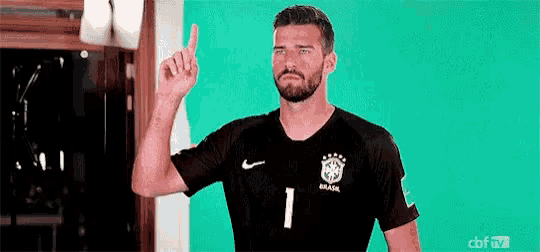 a man with a beard is wearing a black soccer jersey and pointing up with his finger .