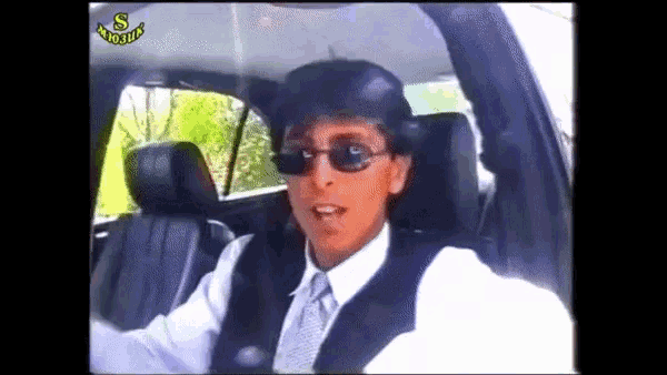 a man wearing sunglasses and a vest is driving a car with the letters s on the bottom left