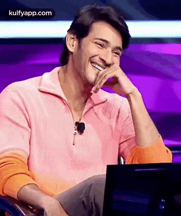 a man in a pink sweater is laughing while sitting in front of a computer .