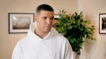a man in a white hoodie is standing in front of a plant in a living room .
