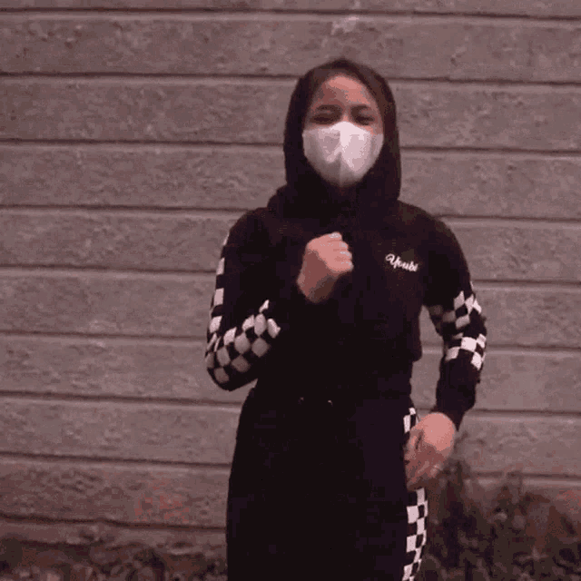 a woman wearing a black and white checkered outfit and a face mask stands in front of a brick wall