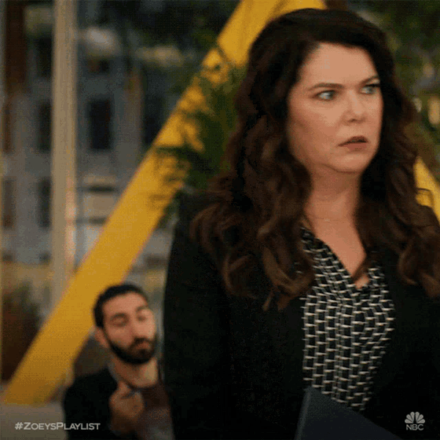 a woman in a black jacket stands in front of a man with a beard and the hashtag #zoesplaylist