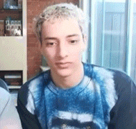 a young man wearing a blue tie dye sweater is sitting in a living room looking at the camera .