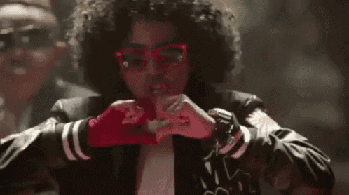 a man with curly hair and glasses is making a heart with his hands .