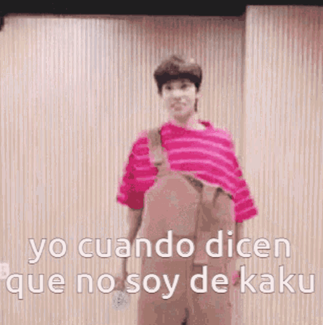 a man in a pink shirt and overalls is standing in front of a wooden wall .