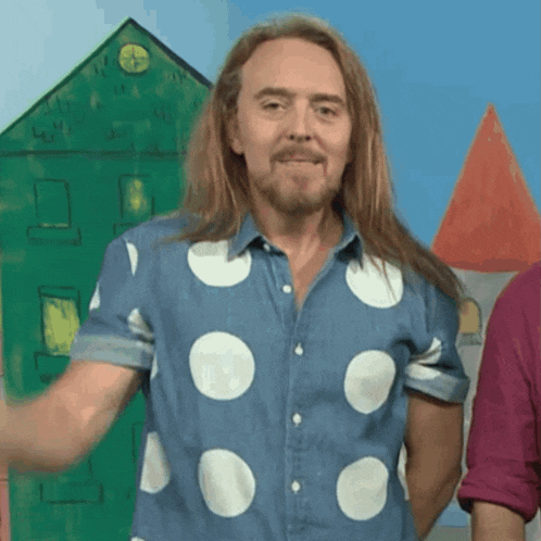 a man with long hair and a polka dot shirt stands in front of a green house