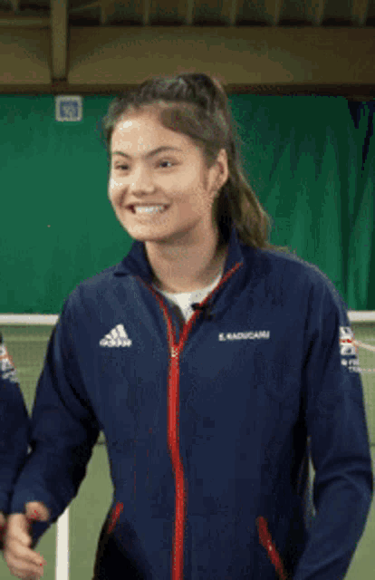a woman wearing an adidas jacket is clapping her hands on a tennis court