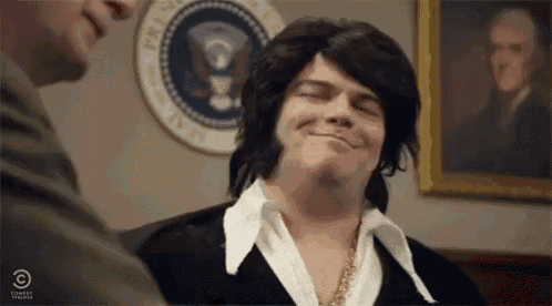 a man wearing a wig and a white shirt is smiling in front of a picture of elvis presley .