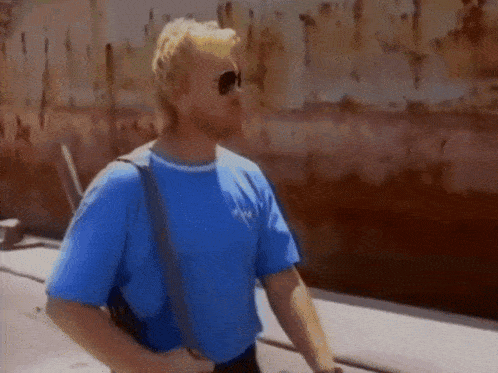 a man wearing sunglasses and a blue shirt is standing in front of a rusty wall .