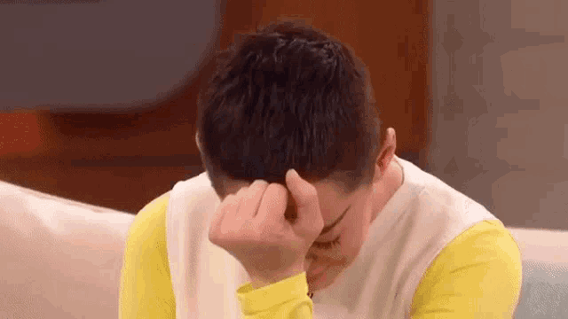 a man in a yellow shirt is sitting on a couch covering his face with his hand .