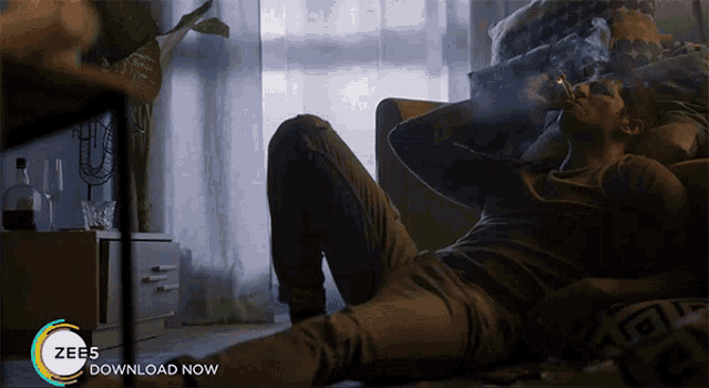 a man is smoking a cigarette while laying on the floor next to a sign that says ' zee5 download now '
