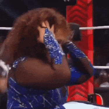 a woman with curly hair is sitting in a wrestling ring with her fists in the air .