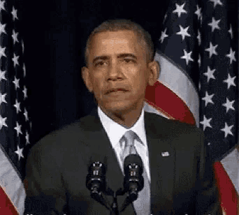 barack obama is giving a speech in front of a microphone in front of american flags .