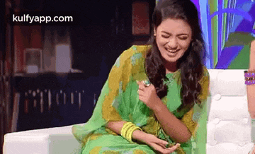 a woman in a green and yellow saree is sitting on a couch laughing .