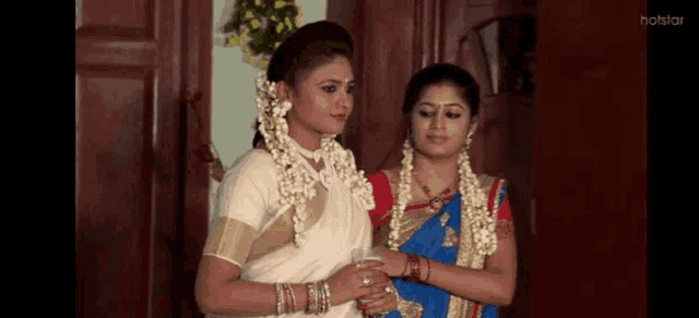 two women are standing next to each other in a room . one of the women is wearing a white saree .