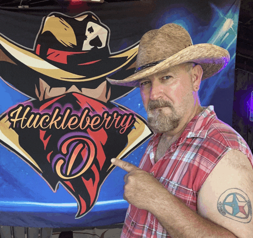 a man wearing a cowboy hat points to his tattoo in front of a huckleberry d. banner