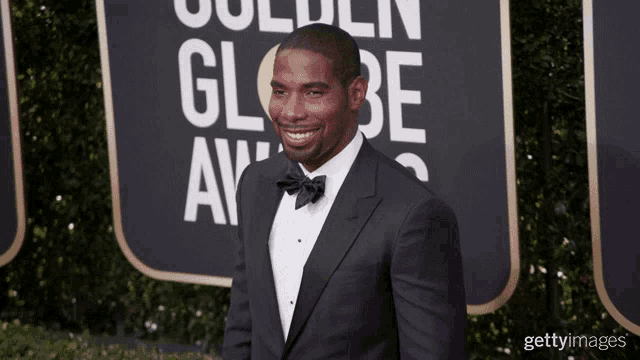 a man in a tuxedo and bow tie smiles on a red carpet in front of a sign that says golden globe awards