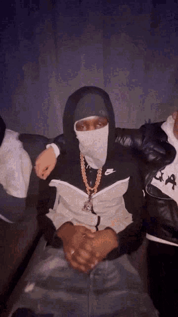 a man wearing a mask and a hoodie is sitting on a couch with his hands folded .
