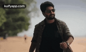 a man with a beard wearing sunglasses and a watch is walking on a beach .