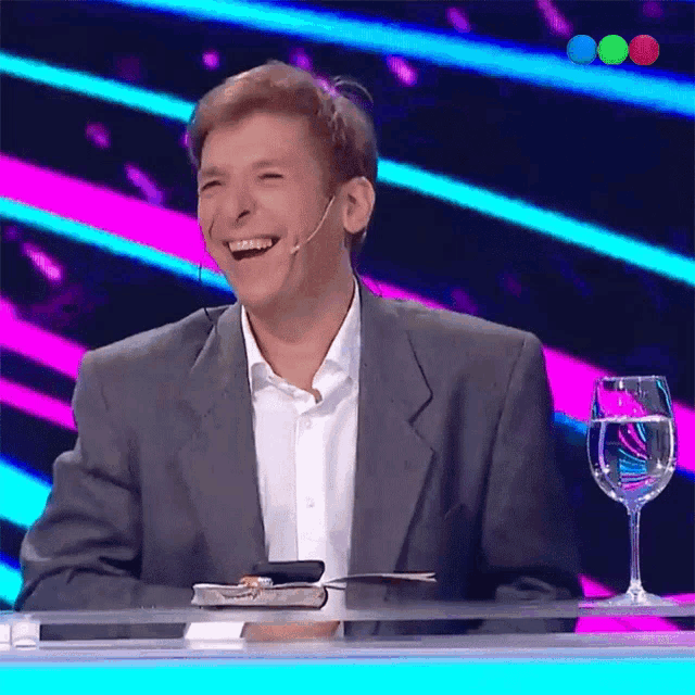 a man in a suit is laughing while sitting at a table with a book and a glass of water
