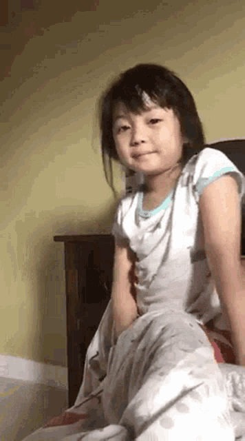 a little girl is sitting on a bed with a blanket on her lap and looking at the camera .