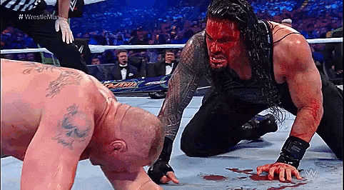 a wrestler with blood on his face is kneeling down next to another wrestler in a ring .