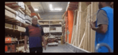 two men are standing in a warehouse talking to each other . one of the men is wearing a blue vest .
