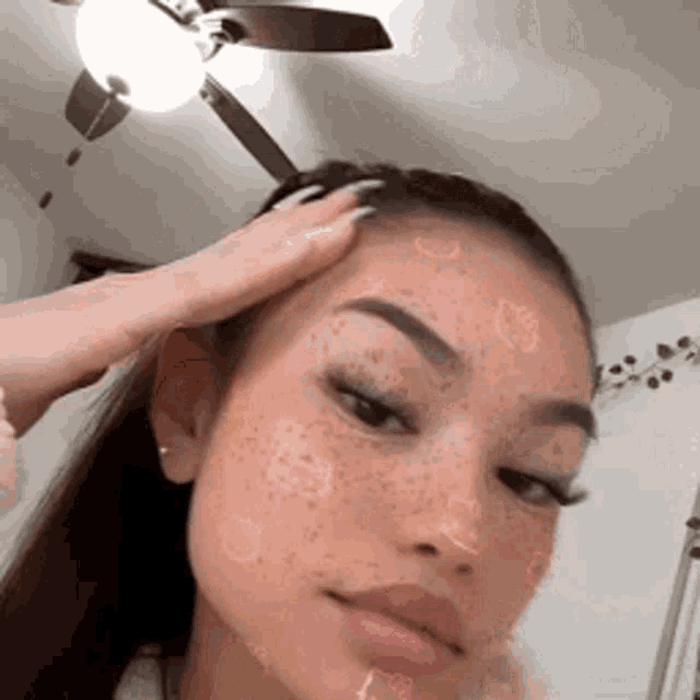 a woman with freckles on her face is touching her head while a ceiling fan is hanging from the ceiling .