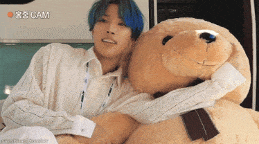a man with blue hair is hugging a large teddy bear with cam written on the bottom