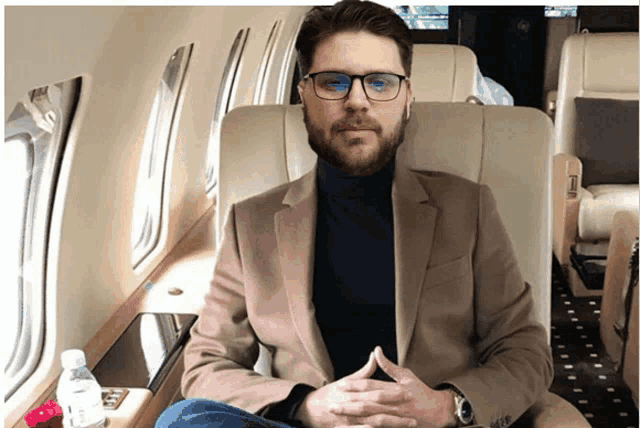 a man in a suit and glasses sits on a plane with his hands folded