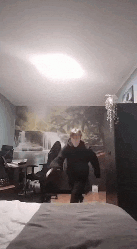 a man in a black hoodie is dancing in a bedroom with a painting on the wall .