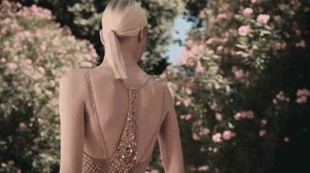 a woman in a dress with a ponytail is standing in front of a bush of pink flowers