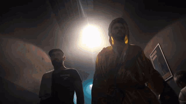 a man in a yellow robe is standing next to another man in a black shirt that says ' bomber ' on it
