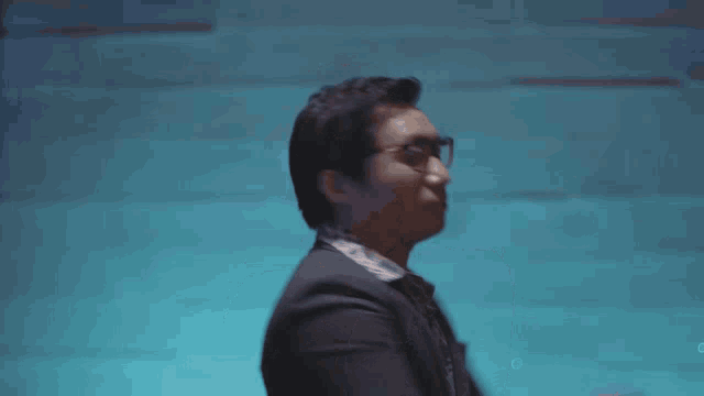 a man wearing glasses and a suit is dancing in front of a blue wall .
