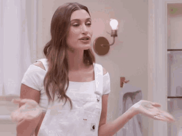a woman in a white top and white overalls is standing in a bathroom with her hands outstretched .