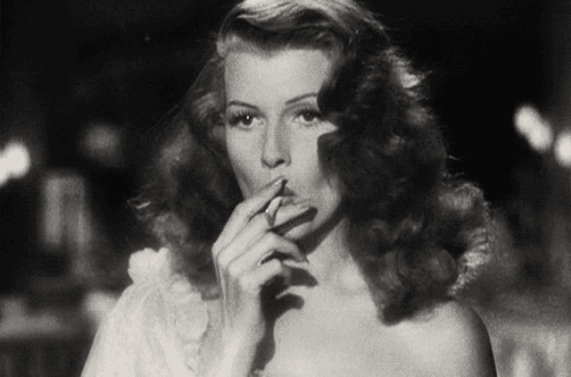 a woman in a white dress is smoking a cigarette in a black and white photo .