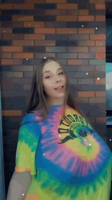 a woman in a colorful tie dye shirt is standing in front of a brick wall and looking at the camera .