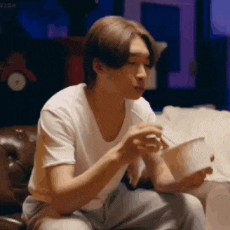 a man in a white shirt is sitting on a couch eating noodles with chopsticks .