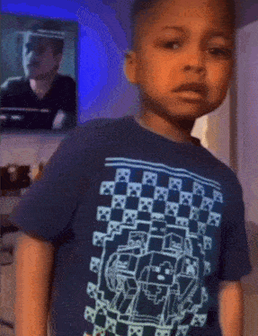 a young boy wearing a blue minecraft shirt is standing in front of a television .