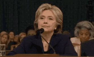 hillary clinton is talking on a cell phone while sitting at a desk in front of a crowd .